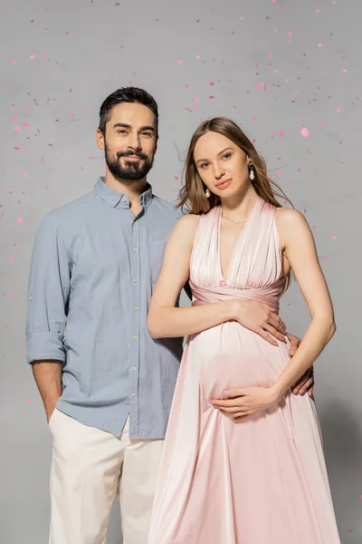 Portrait of smiling man hugging elegant and pregnant wife in pink dress while standing under confetti during baby shower party on grey background, expecting parents concept — Stock Photo