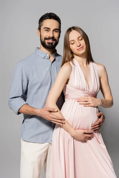 Позитивный мужчина обнимает элегантную и беременную жену в розовом платье и смотрит в камеру, стоя вместе на сером фоне, ожидая концепции родителей, новых начинаний — стоковое фото