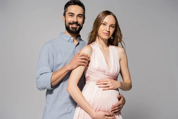 Portrait of positive and bearded man hugging elegant pregnant wife in pink dress while standing together isolated on grey, expecting parents concept, new beginnings, excitement — Stock Photo