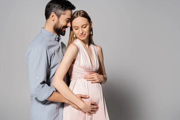 Homem feliz e barbudo abraçando a mulher grávida em vestido elegante enquanto estão juntos em fundo cinza, esperando o conceito dos pais, marido e esposa — Fotografia de Stock
