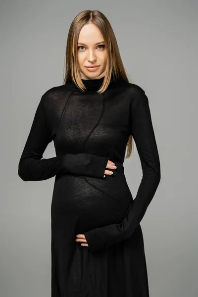 Trendy and fair haired expecting mother wearing stylish black dress touching belly and looking at camera while posing isolated on grey, growing new life concept — Stock Photo