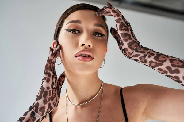 Portrait of young asian woman with brunette hair, bold makeup and expressive gaze posing in animal print gloves and silver accessories while looking at camera on grey background, trendy spring — Stock Photo