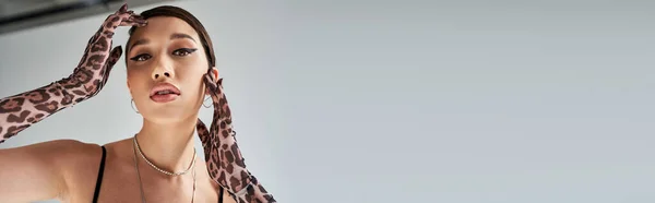 Moda primaverile, ritratto di attraente donna asiatica con trucco audace, in collane d'argento, orecchini e guanti di stampa animalier guardando la fotocamera su sfondo grigio, banner — Foto stock