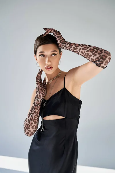 Appealing asian woman with brunette hair and bold makeup posing in black strap dress and animal print gloves on grey background, spring style, modern fashion photography — Stock Photo