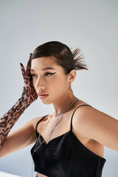 Sensual asian fashion model with bold makeup and trendy hairstyle, wearing black strap dress, animal print glove and silver accessories, holding hand near face on grey background, stylish spring — Stock Photo
