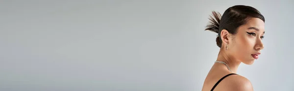 Retrato de perfil de mujer asiática joven con cabello moreno, maquillaje audaz, peinado de moda posando en collares de plata y mirando hacia otro lado sobre fondo gris, primavera elegante, fotografía de moda, pancarta - foto de stock