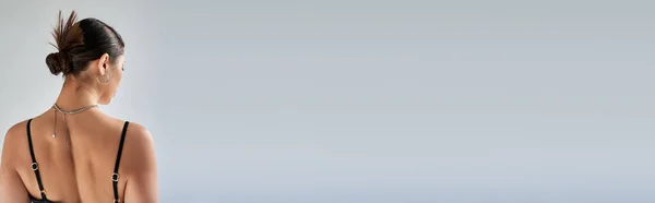 Vista posteriore di giovane donna bruna in abito alla moda primavera, acconciatura alla moda, abito cinturino nero, collane d'argento, in piedi su sfondo grigio, generazione z, banner — Foto stock