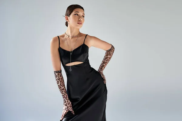 Spring style, expressive asian woman with brunette hair and bold makeup standing with hand on hip in black elegant strap dress, animal print gloves and silver necklaces on grey background — Stock Photo