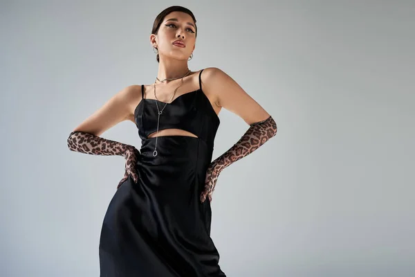 Vista de ángulo bajo de morena mujer asiática con maquillaje audaz posando con las manos en las caderas sobre fondo gris, vestido de correa elegante negro, guantes de impresión animal, accesorios de plata, fotografía de moda de primavera — Stock Photo