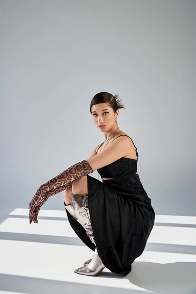 Full length of young asian woman in elegant spring outfit posing on haunches on grey background with lighting and shadows, black strap dress, animal print gloves, silver boots, generation z — Stock Photo