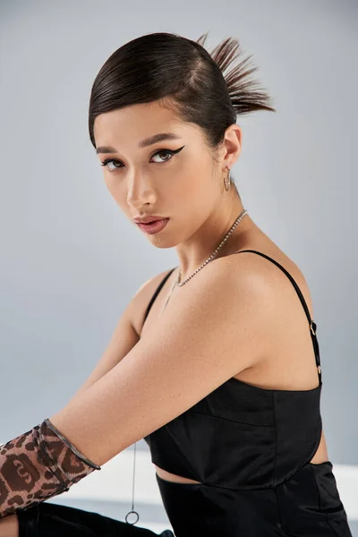 Spring style asian woman with bold makeup, brunette hair and expressive gaze looking at camera on grey background, black strap dress, silver necklaces, animal print glove, fashion shoot — Stock Photo