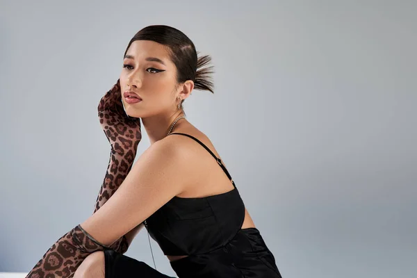 Charming asian woman with trendy hairstyle and bold makeup posing in stylish spring outfit on grey background, black strap dress, animal print gloves, fashion photography, generation z — Stock Photo