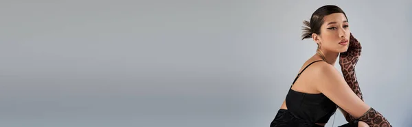 Mulher asiática na moda em roupa de primavera, com cabelo morena e penteado na moda posando em vestido de cinta preta e luvas de impressão animal enquanto olha para o fundo cinza, fotografia de moda, banner — Fotografia de Stock