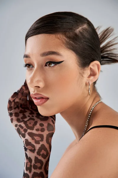 Portrait of mesmerizing asian woman with brunette hair, bold makeup, trendy hairstyle, silver necklaces and animal print glove posing with hand near face on grey background, spring fashion — Stock Photo