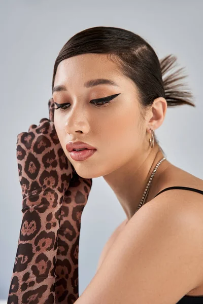 Portrait of beautiful and dreamy asian woman in animal print gloves and silver accessories, with trendy hairstyle and bold makeup on grey background, stylish spring, fashion shoot — Stock Photo