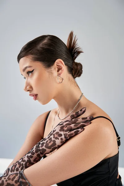 Retrato de mujer asiática joven con peinado de moda y maquillaje audaz posando en guantes de impresión animal, vestido de correa negra y accesorios de plata sobre fondo gris, concepto de moda de primavera - foto de stock