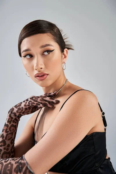 Портрет модной азиатской женщины с модной прической и смелым макияжем, позирующим в черном платье, перчатками для печати животных и серебряными ожерельями, отводя взгляд на сером фоне, стильная весна — стоковое фото