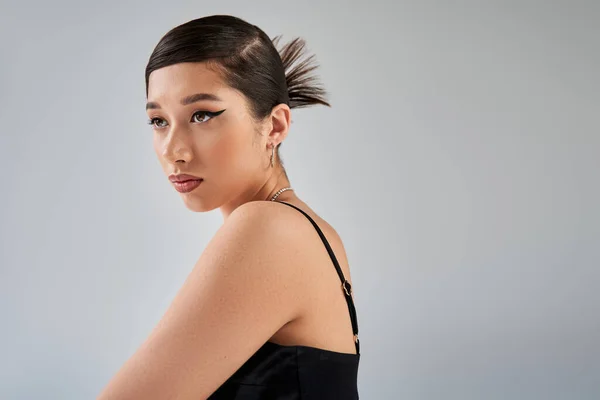 Spring fashion photography, portrait of stylish asian model in black strap dress, with trendy hairstyle, bold makeup and expressive gaze looking away on grey background, generation z — Stock Photo