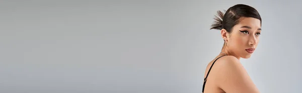 Ritratto di ipnotizzante donna asiatica con acconciatura alla moda, capelli castani e trucco audace guardando altrove mentre posa su sfondo grigio con spazio copia, fotografia di moda primaverile, banner — Foto stock