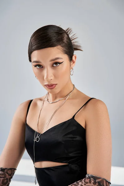 Portrait of young asian woman with brunette hair, bold makeup and seductive gaze posing in black strap dress and silver accessories on grey background, stylish spring, fashion shoot — Stock Photo