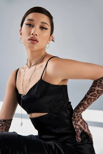 Printemps à la mode, portrait de femme asiatique sensuelle au regard expressif et au maquillage lumineux, en robe à bretelles noires et accessoires argentés, tenant la main sur la hanche sur fond gris avec éclairage et ombres — Photo de stock