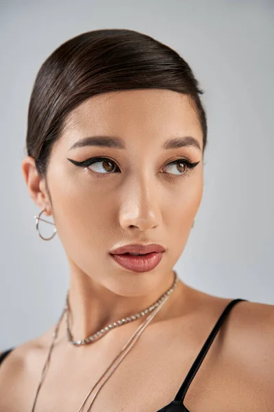 Portrait of appealing and young asian woman in silver accessories, with brunette hair, bold makeup and trendy hairstyle looking away on grey background, stylish spring, fashion shoot — Stock Photo