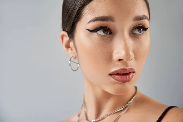 Portrait of asian woman in silver necklaces and earring, with brunette hair, bold makeup and expressive gaze looking away on grey background, trendy spring, fashion photography — Stock Photo
