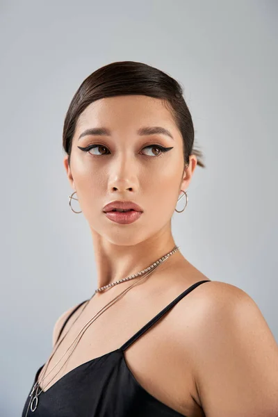 Portrait de femme asiatique séduisante en élégante robe noire, colliers et boucles d'oreilles en argent, avec maquillage audacieux et cheveux bruns regardant loin sur fond gris, printemps à la mode — Photo de stock