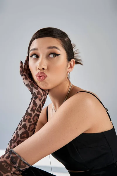 Portrait of fashionable asian woman with bold makeup and brunette hair pouting lips and looking away on grey background, spring fashion, black dress, animal print gloves, silver accessories — Stock Photo