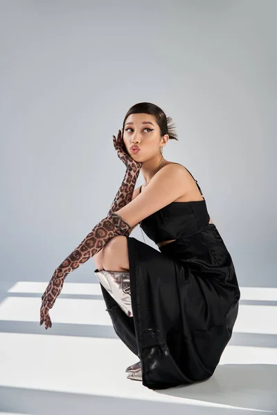 Attractive asian woman in black elegant dress, animal print gloves and silver boots sitting on haunches, blowing kiss and looking away on grey background with lighting, spring fashion photography — Stock Photo