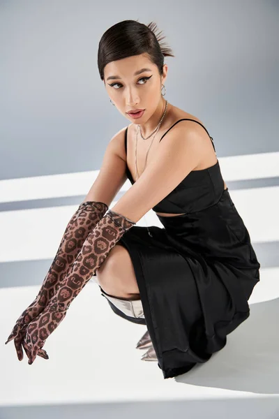 Full length of young asian woman in black elegant dress, animal print gloves and silver boots posing on haunches and looking away on grey background with lighting, high fashion, spring style — Stock Photo