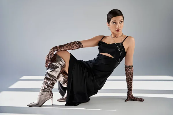 Full length of trendy asian woman with bold makeup, in silver boots, black strap dress and animal print gloves posing and looking away on grey background with lighting and shadows, spring fashion — Stock Photo