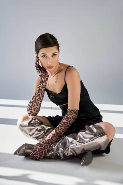 Primavera de moda, mujer asiática joven con maquillaje audaz y mirada expresiva, en vestido de correa negro, guantes de impresión animal y botas de plata sentado y mirando a la cámara sobre fondo gris con iluminación - foto de stock