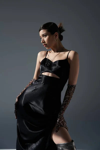 Concepto de primavera de moda, mujer asiática joven en vestido de correa negro y guantes de impresión animal posando con la mano en la cadera y mirando hacia otro lado sobre fondo gris oscuro - foto de stock