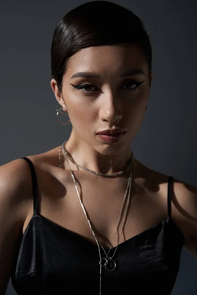 Портрет очаровательной азиатской женщины с смелым макияжем и брюнеткой волосы, в черном платье ремешок и серебряные ожерелья глядя на камеру на темно-сером фоне, поколение z, весенний стиль концепции — стоковое фото