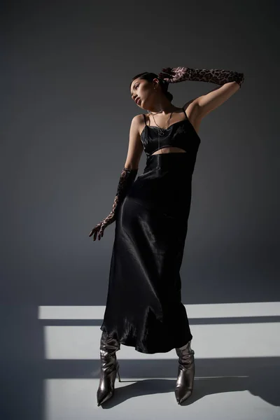 Повна довжина витонченої азіатки в чорній сукні з ремінцем, рукавички для тварин та срібні черевики, що стоять у стильній позі на темно-сірому тлі з освітленням, весняна модна фотографія — стокове фото