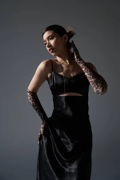 Юношеская мода, стильная весна, чувственная азиатская женщина с брюнетками, в черном элегантном платье и перчатках для печати животных, позирующая с рукой на бедре на темно-сером фоне — стоковое фото