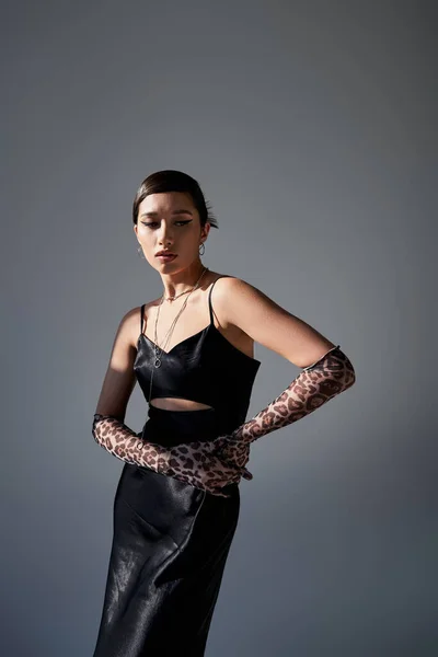 Young and fashionable asian woman with bold makeup, brunette hair, in black strap dress and animal print gloves standing with hands on hip on dark grey background, spring fashion concept — Stock Photo