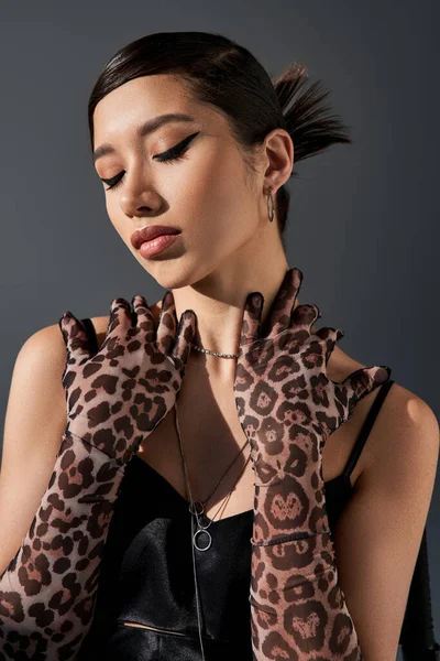 Portrait de femme asiatique sensuelle avec un maquillage audacieux, en robe à bretelles noires et gants à imprimé animal posant les yeux fermés sur fond gris foncé, concept printemps tendance, génération z — Photo de stock