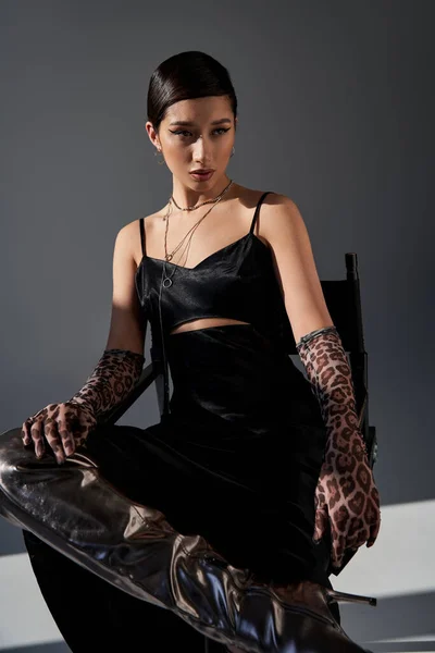 Spring fashion concept, young asian woman with bold makeup, brunette hair, black strap dress, animal print gloves and silver boot sitting on chair on grey background with lighting, generation z — Stock Photo