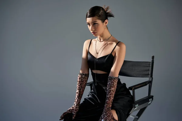 Молодая азиатская модель с брюнеткой волосы позируют на стуле в модном весеннем наряде, черный ремень платье, перчатки для печати животных, серебряные ожерелья на сером фоне с освещением, поколение z — стоковое фото