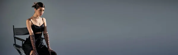 Moda primavera, expressiva mulher asiática com cabelo morena sentado na cadeira em vestido de cinta preta, luvas de impressão animal e olhando para o fundo cinza com iluminação, fotografia de moda, banner — Fotografia de Stock