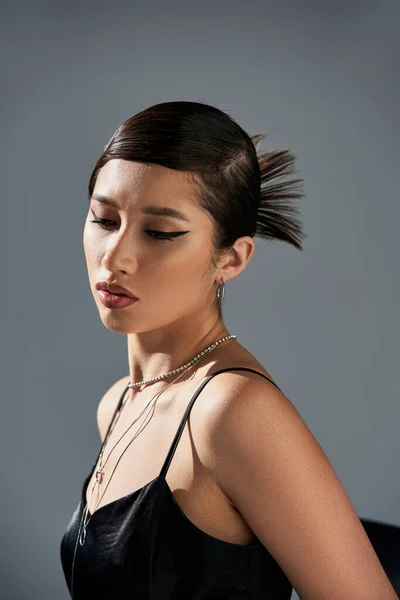 Portrait de femme asiatique séduisante aux cheveux bruns, maquillage audacieux, coiffure tendance, robe à bretelles noires et accessoires argentés posant dans l'éclairage sur fond gris, concept de mode printanière — Photo de stock