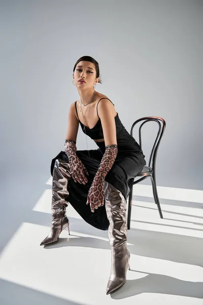 Full length of sensual asian woman with expressive gaze wearing black strap dress, animal print gloves and silver boots while sitting on chair on grey background with lighting, fashionable spring — Stock Photo