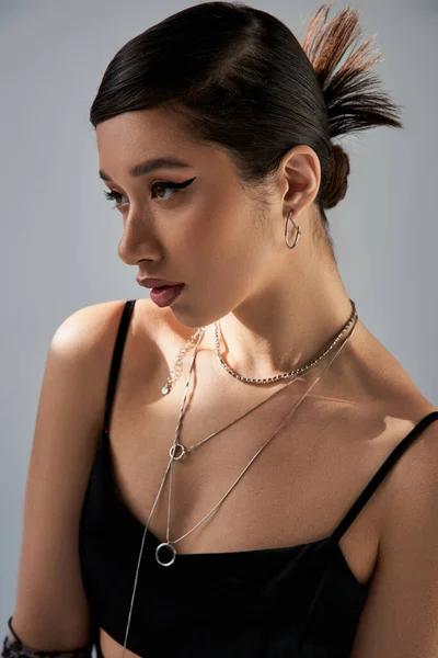 Портрет брюнетки азиатской женщины с смелым макияжем, модной прической и выразительным взглядом, в черном платье с ремешком и серебряными ожерельями, смотрящими в сторону на сером фоне с освещением, модной весенней концепцией — стоковое фото
