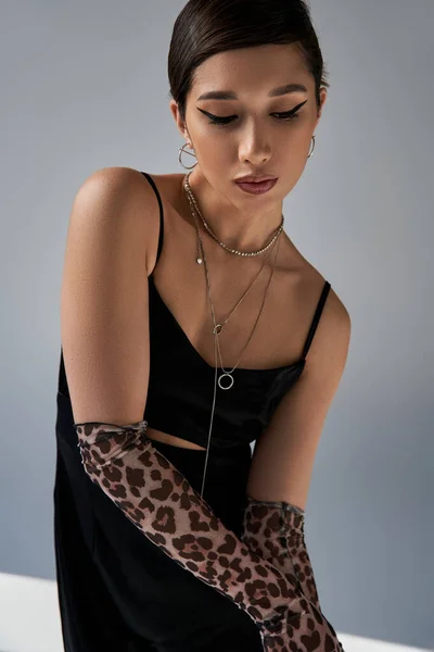 Fashionable asian woman with bold makeup, brunette hair, silver necklaces and animal print gloves posing in black strap dress on grey background, stylish spring, generation z — Stock Photo
