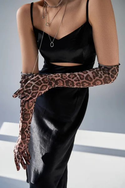 Partial view of fashionable and elegant woman in black dress, silver accessories and animal print gloves standing on grey background with lighting, spring fashion photography — Stock Photo