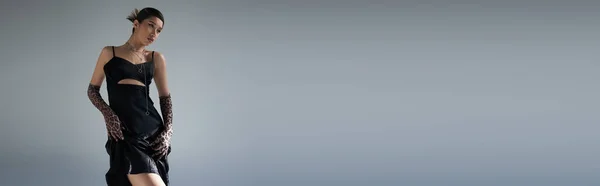 Jovem e sensual asiático mulher com morena cabelo posando em preto vestido cinta e animal imprimir luvas enquanto olhando para o fundo cinza, moderno primavera conceito, geração z, banner — Fotografia de Stock