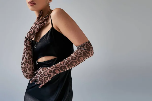 Partial view of fashionable woman in black and elegant strap dress, silver necklaces and animal print gloves touching neck on grey background, trendy spring, generation z — Stock Photo