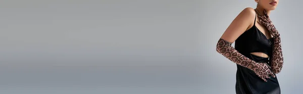 Corte vista de mulher elegante em luvas de impressão animal e vestido de cinta preta segurando a mão perto do pescoço enquanto posando em fundo cinza, conceito de primavera na moda, empoderado, banner — Fotografia de Stock